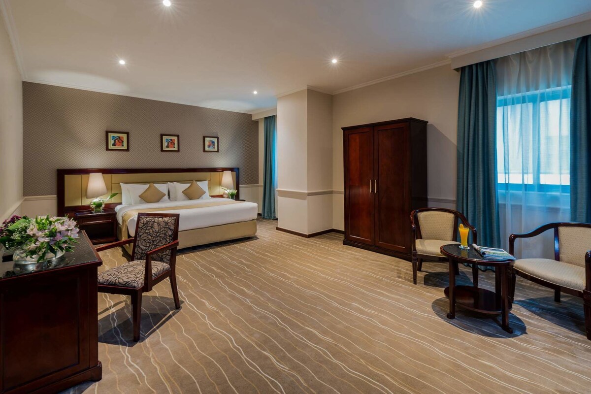 A hotel room with a bed, two chairs, a desk, a wardrobe, and a large window with curtains.