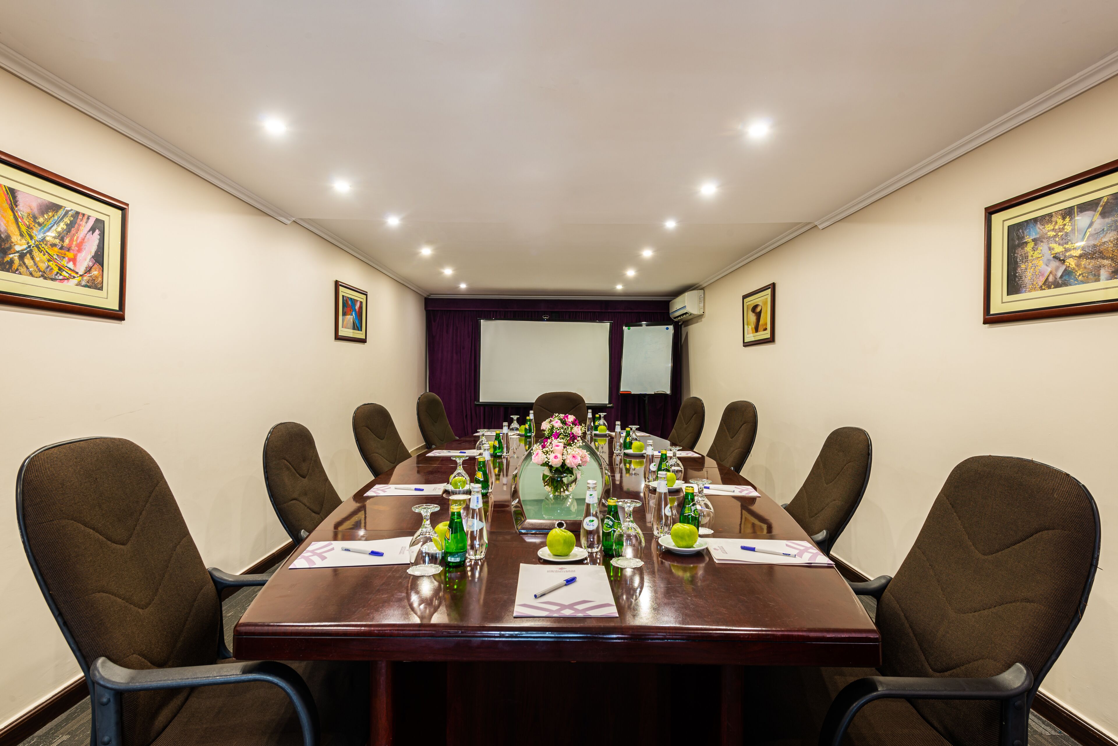 A conference room with a long table, chairs, a projector screen, and framed artwork on the walls.