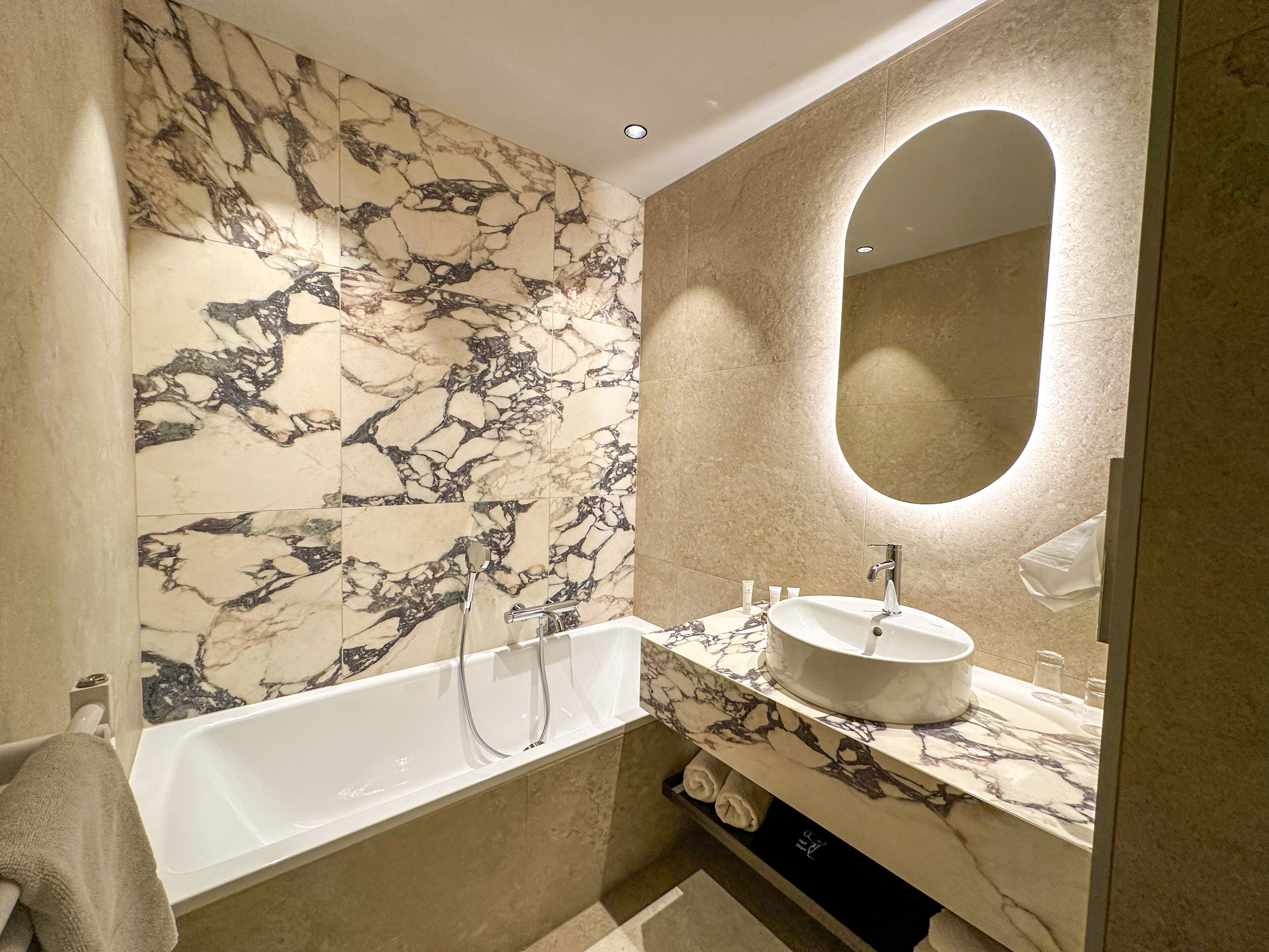 Un baño moderno con pared de mármol, una bañera independiente, un lavamanos con encimera de mármol y un espejo grande.
