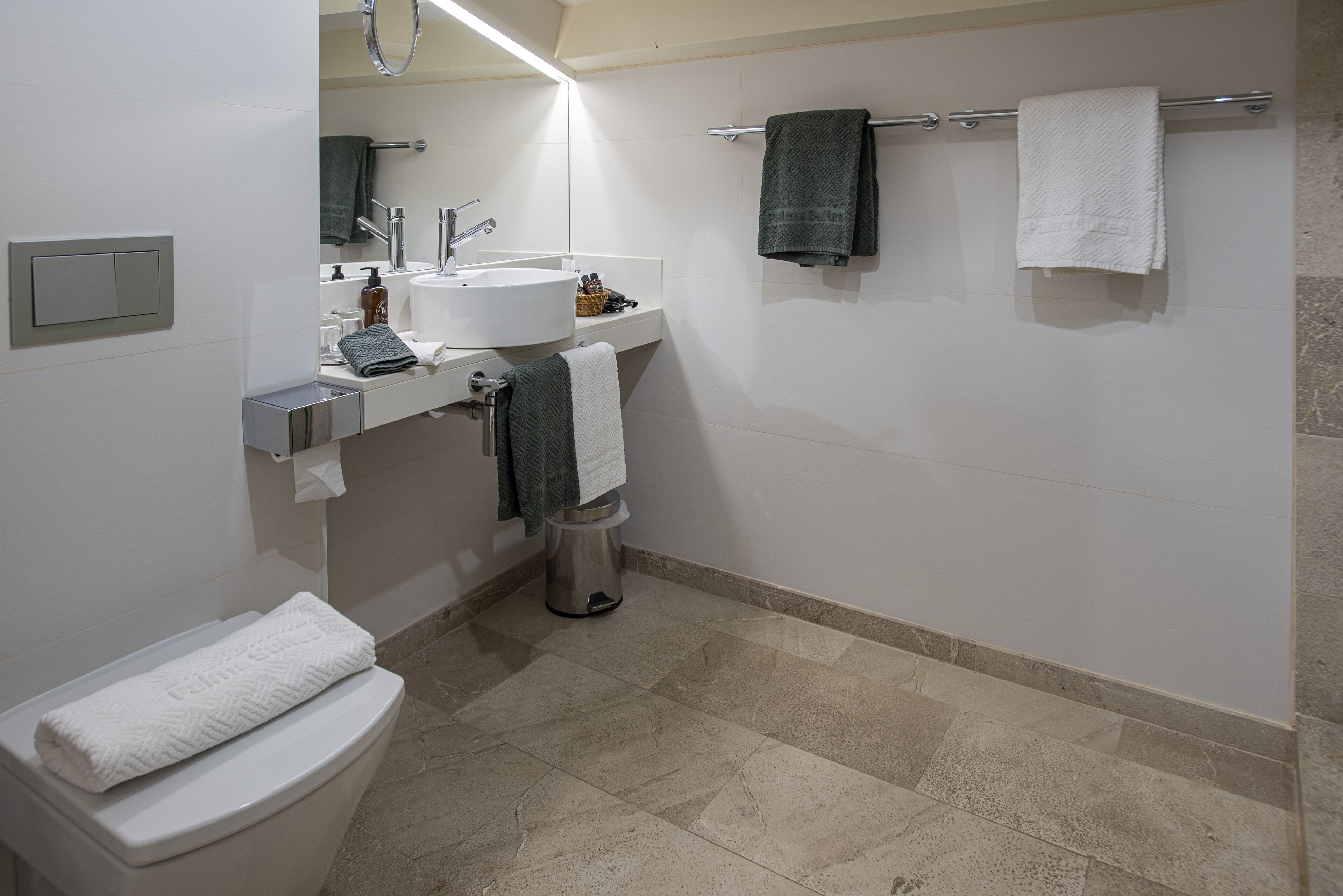 A modern bathroom with a toilet, sink, and mirror. There are towels on the wall and a trash can.