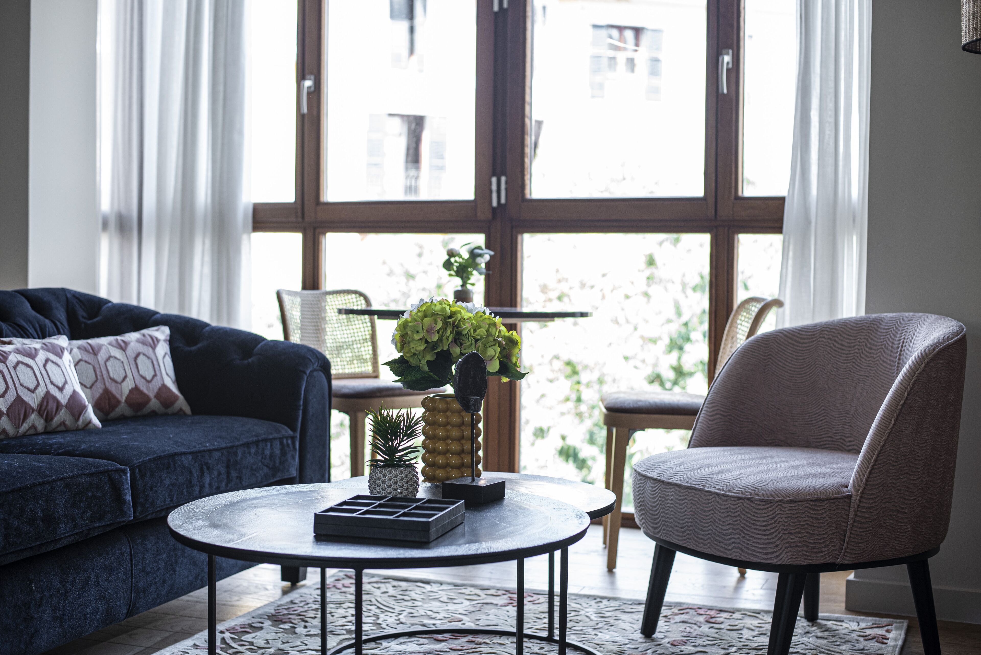 A living room with a dark blue sofa, a round coffee table, and a wicker chair. There is a window with curtains, a small table with a plant, and a decorative pineapple.