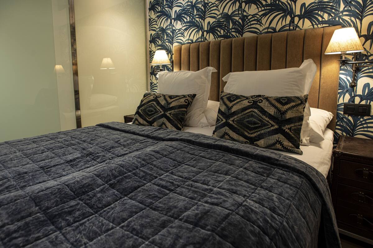 Ein ordentlich bezogenes Bett mit einem gemusterten Quilt, zwei Kissen mit geometrischen Mustern, einem Kopfteil und einem Nachttisch mit Lampe.