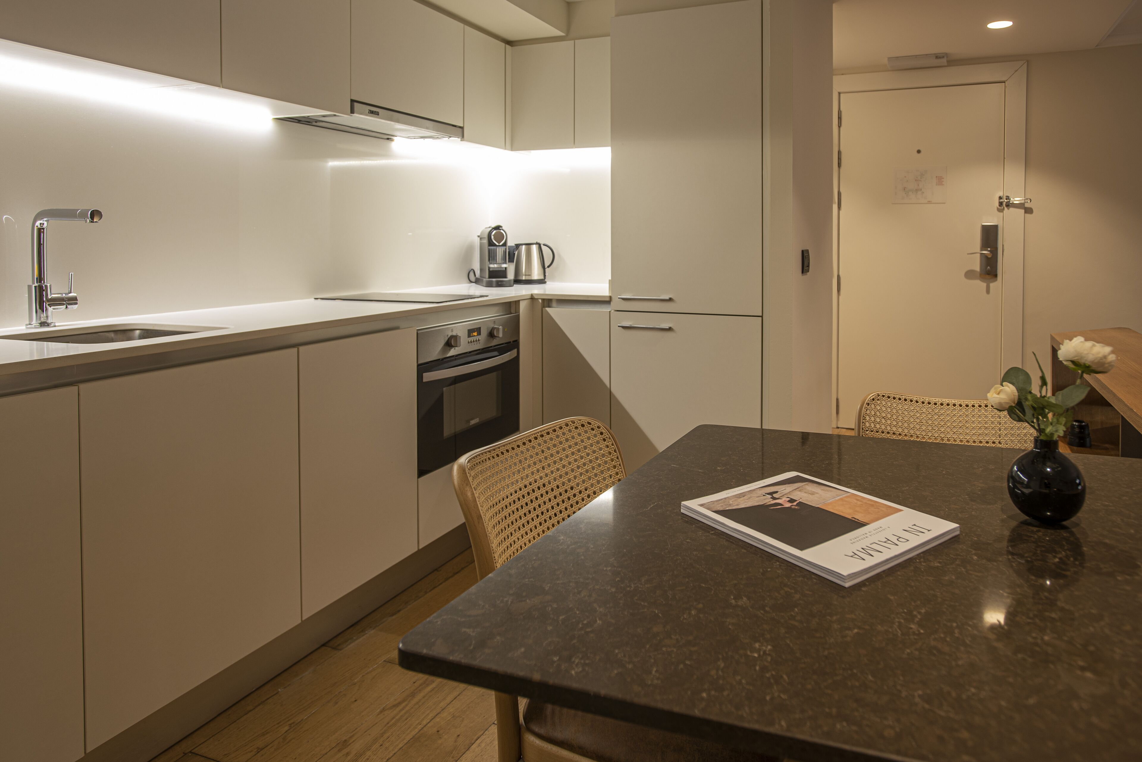 Een moderne keuken met een inbouwoven, een koffiezetapparaat, en een eethoek met een tafel en stoelen.