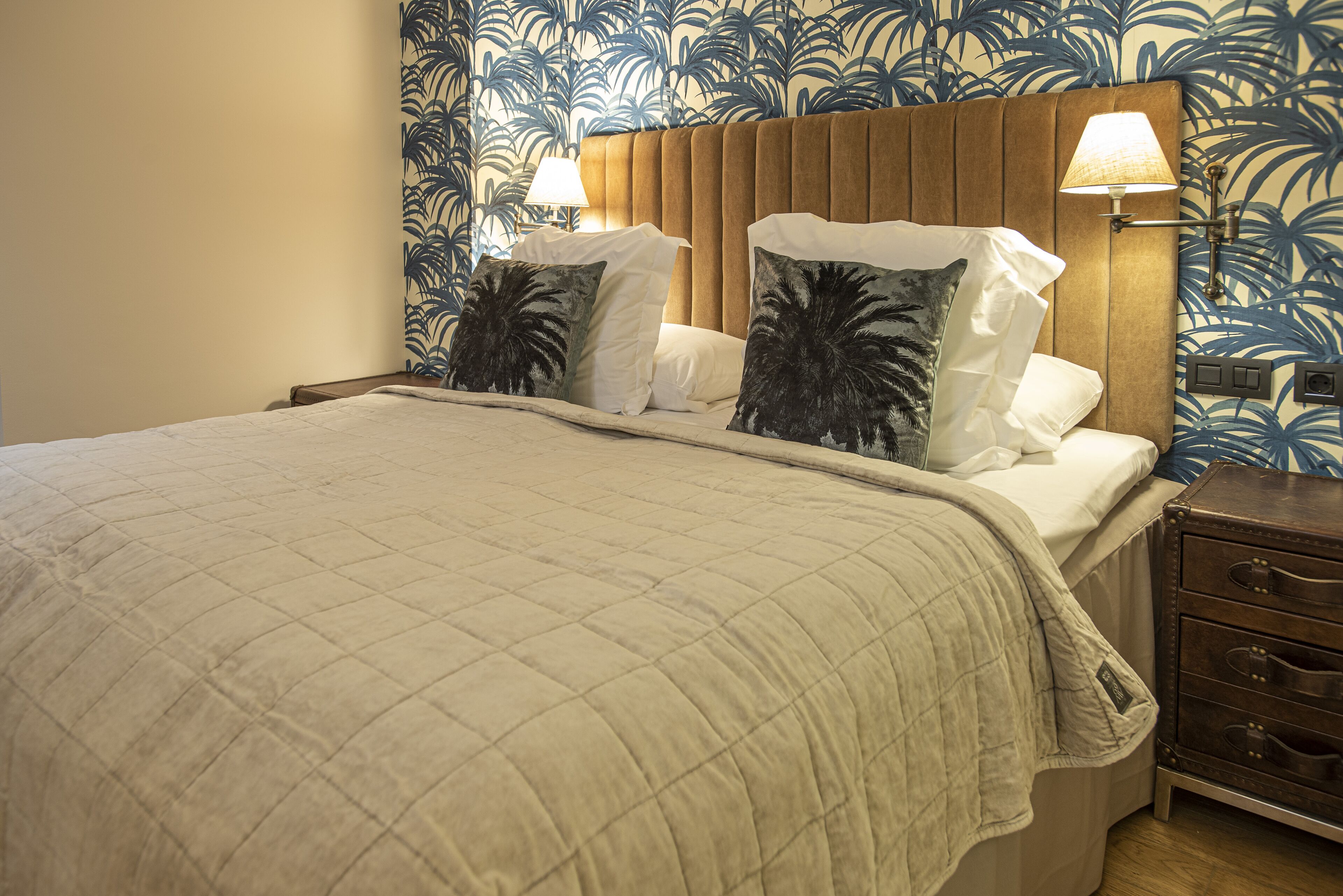 Un lit bien fait, recouvert d’une courtepointe matelassée, avec deux oreillers ornés de motifs de feuilles de palmier, et une tête de lit agrémentée d’une lampe de chaque côté.