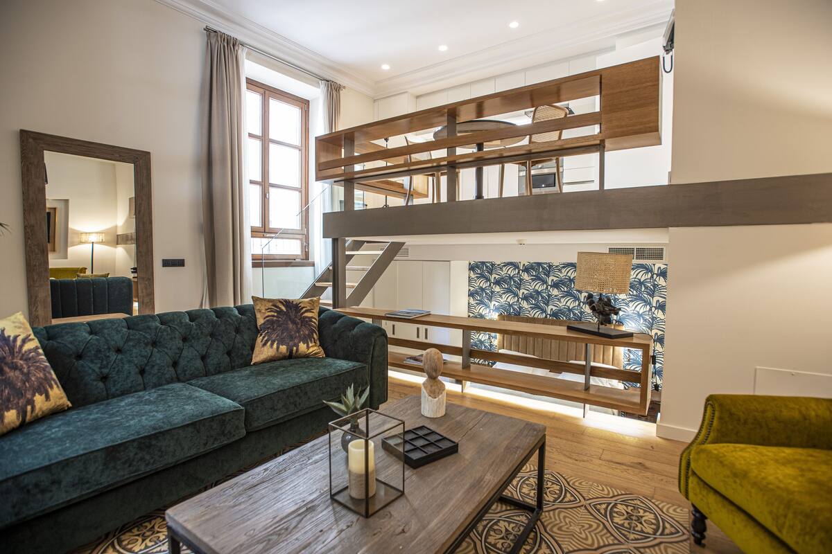 Ein modernes Wohnzimmer mit einem grünen Sofa, einem Holztisch und einem gelben Sessel. Eine Treppe führt zu einer oberen Ebene mit einer Holzwand.