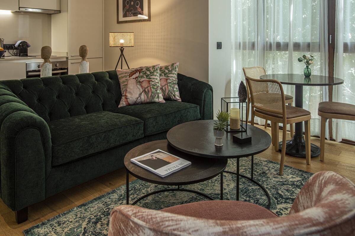 Ein modernes Wohnzimmer mit einem dunkelgrünen Sofa, einem runden Couchtisch und einem Essbereich mit einem Glastisch und Korbstühlen.