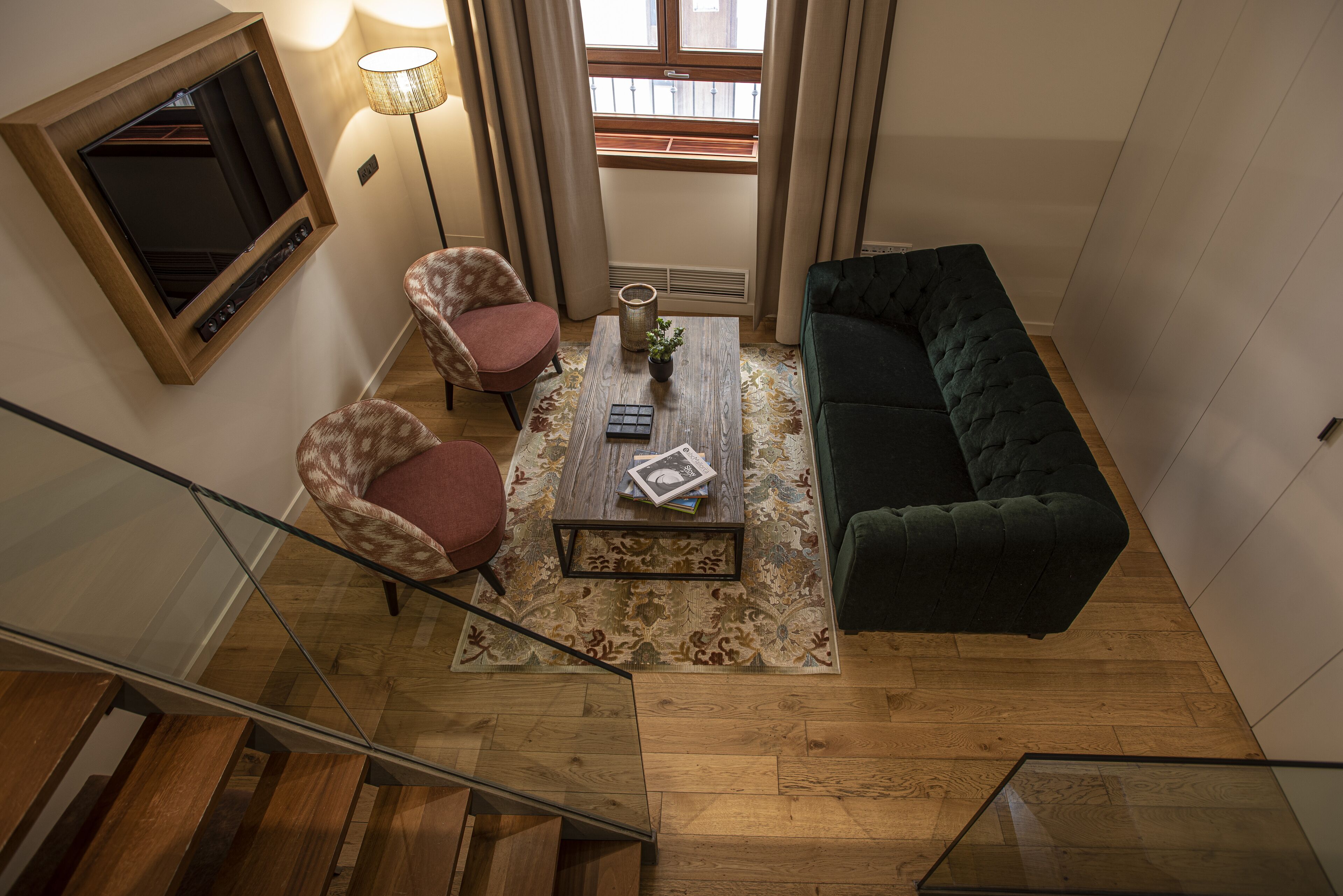 A modern living room with a staircase, a green sofa, two patterned armchairs, a coffee table with books, and a TV mounted on the wall.