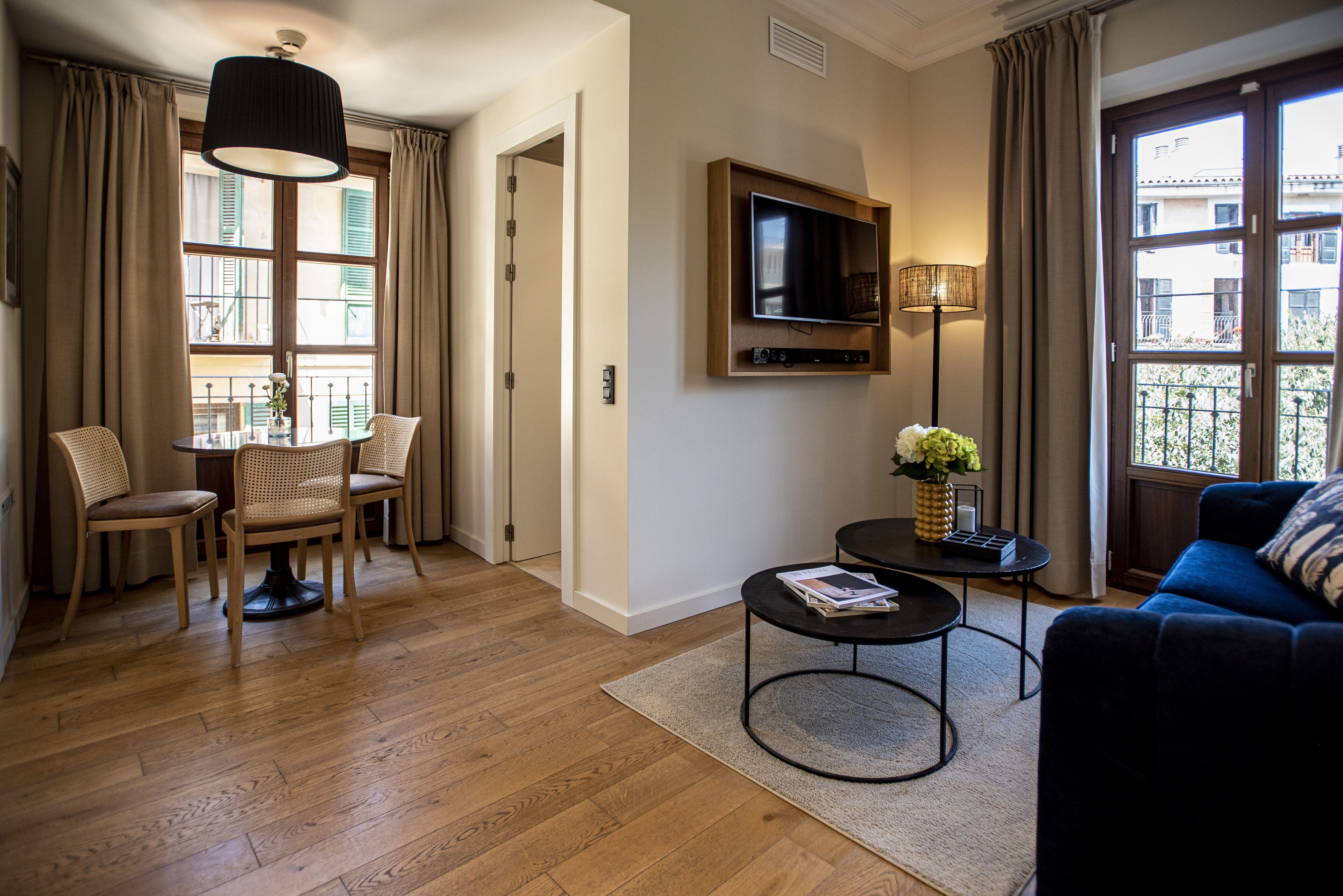 A living room with a sofa, a TV mounted on the wall, a dining table with chairs, and a window with curtains.