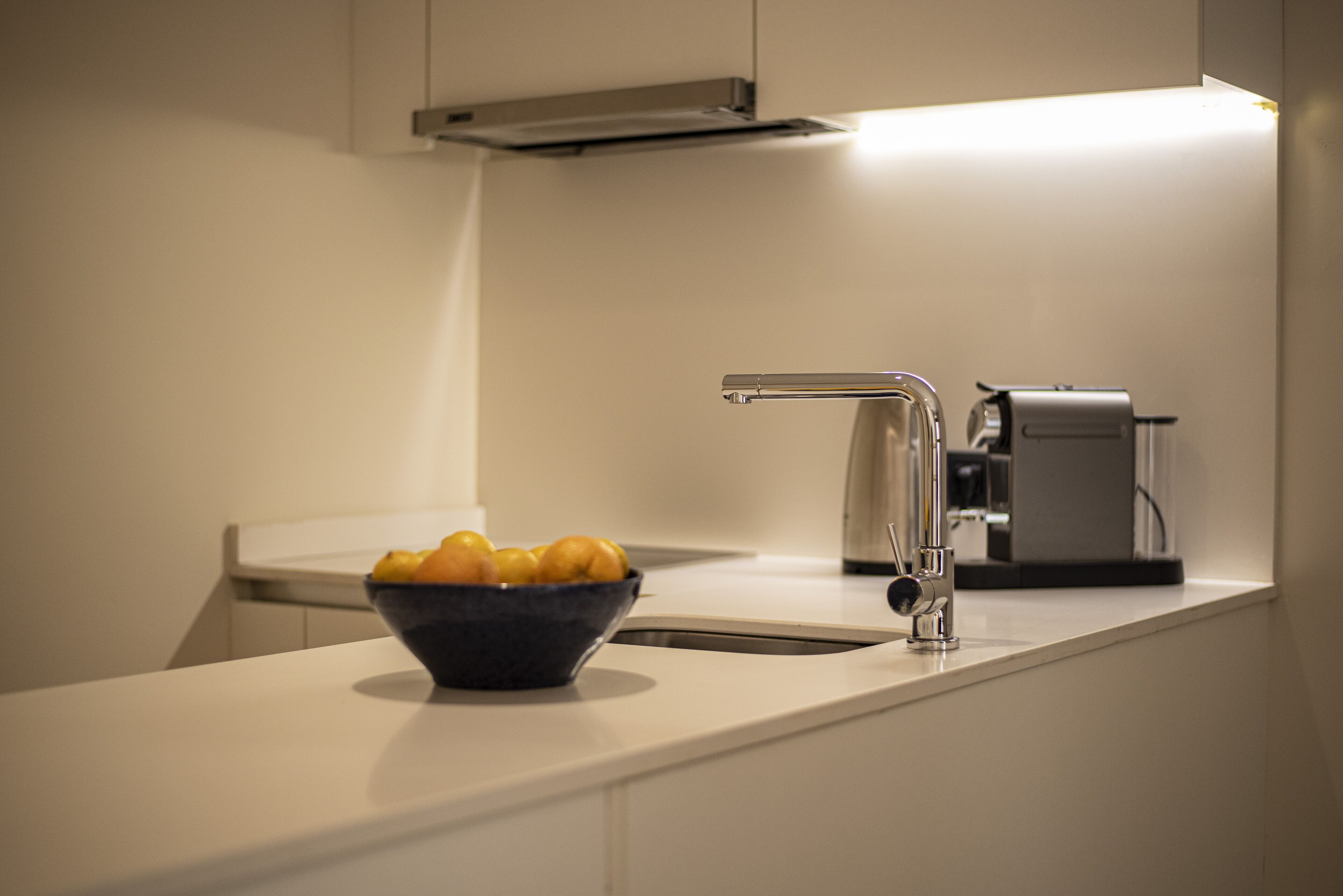 A modern kitchen with a white countertop, a stainless steel sink, and a bowl of fruit.