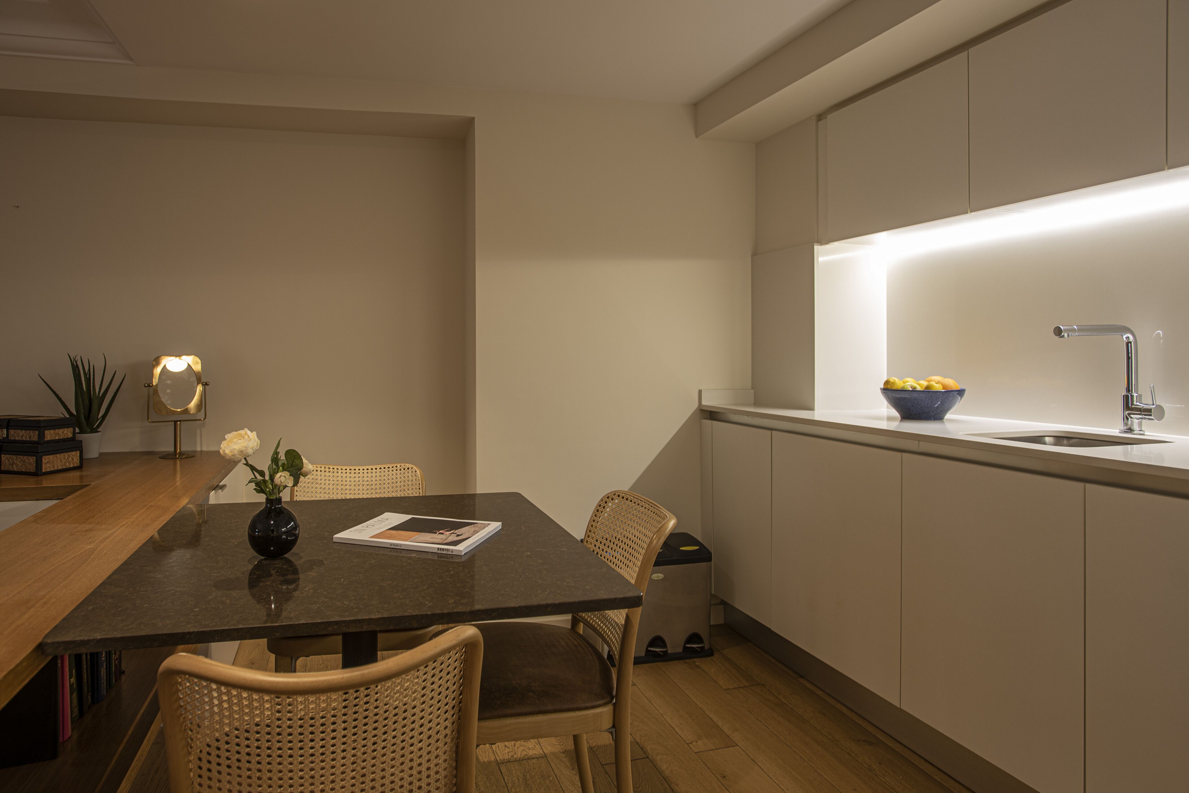 A modern kitchen with a dining area. It includes a dark wooden table with chairs, a small vase with flowers, and a bowl of fruit on the counter.