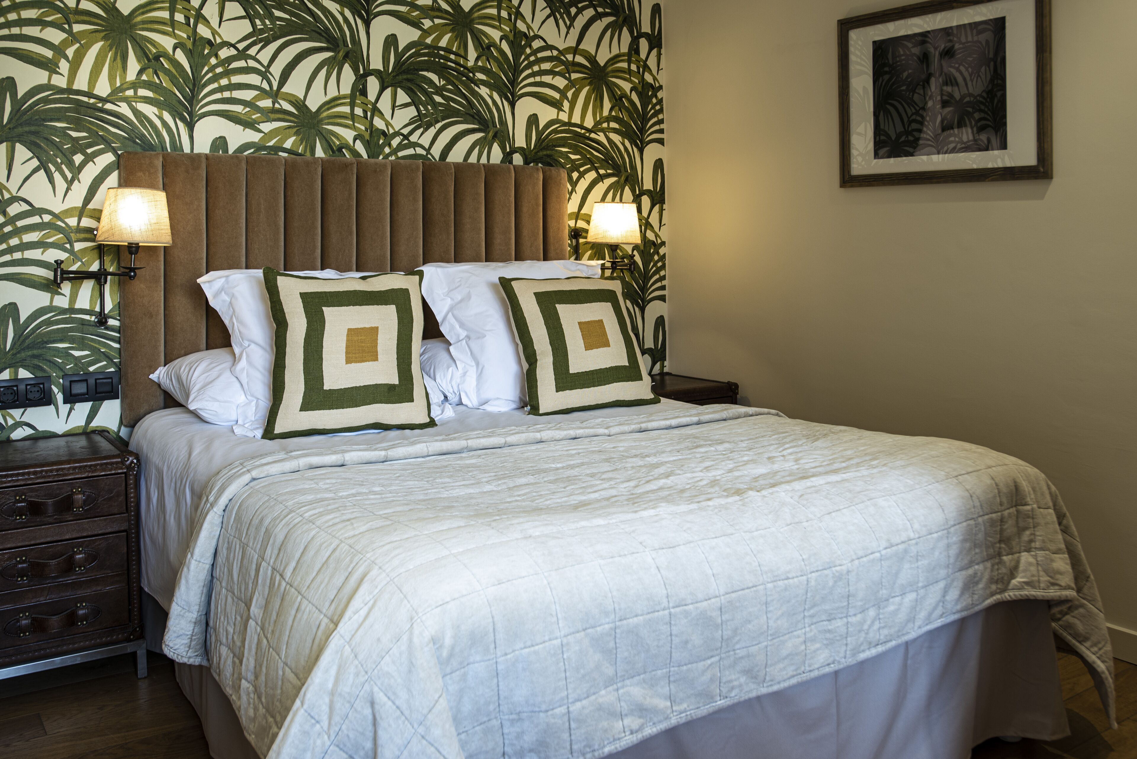 Een net opgemaakt bed met een geborduurde witte dekbedovertrek en twee decoratieve kussens, met aan weerszijden wandlampen.