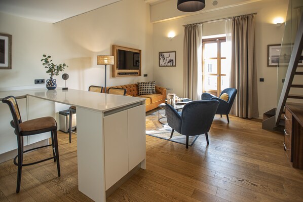 Una cocina moderna con una isla central, un comedor con sofá y sillones, y una escalera que conduce a un nivel superior.