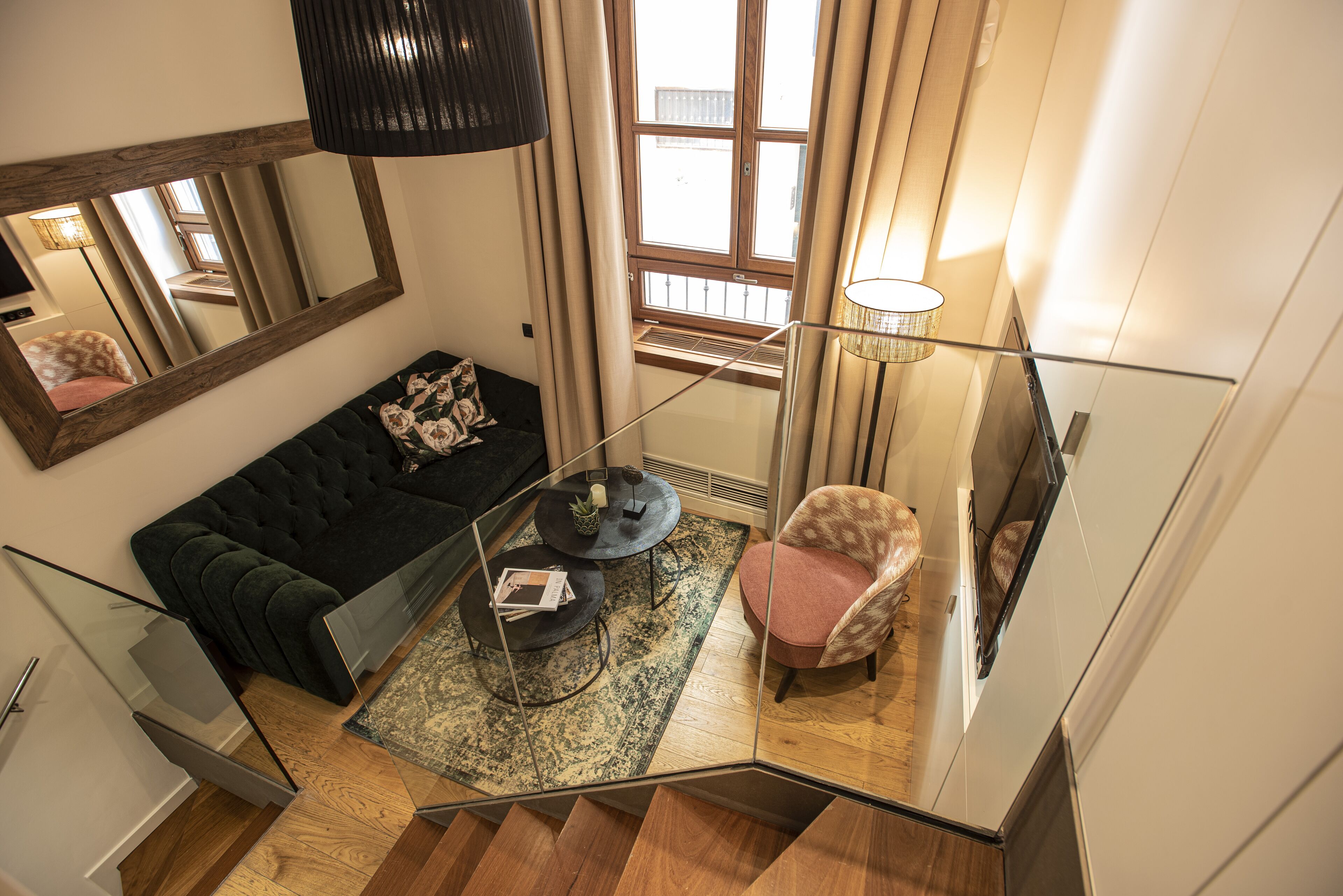 Un salon moderne avec un canapé vert foncé, une table basse ronde et un tapis à motifs. Une chaise avec un coussin rose est placée près d’une fenêtre ornée de rideaux. Un grand miroir est accroché au mur et reflète une partie de la pièce.