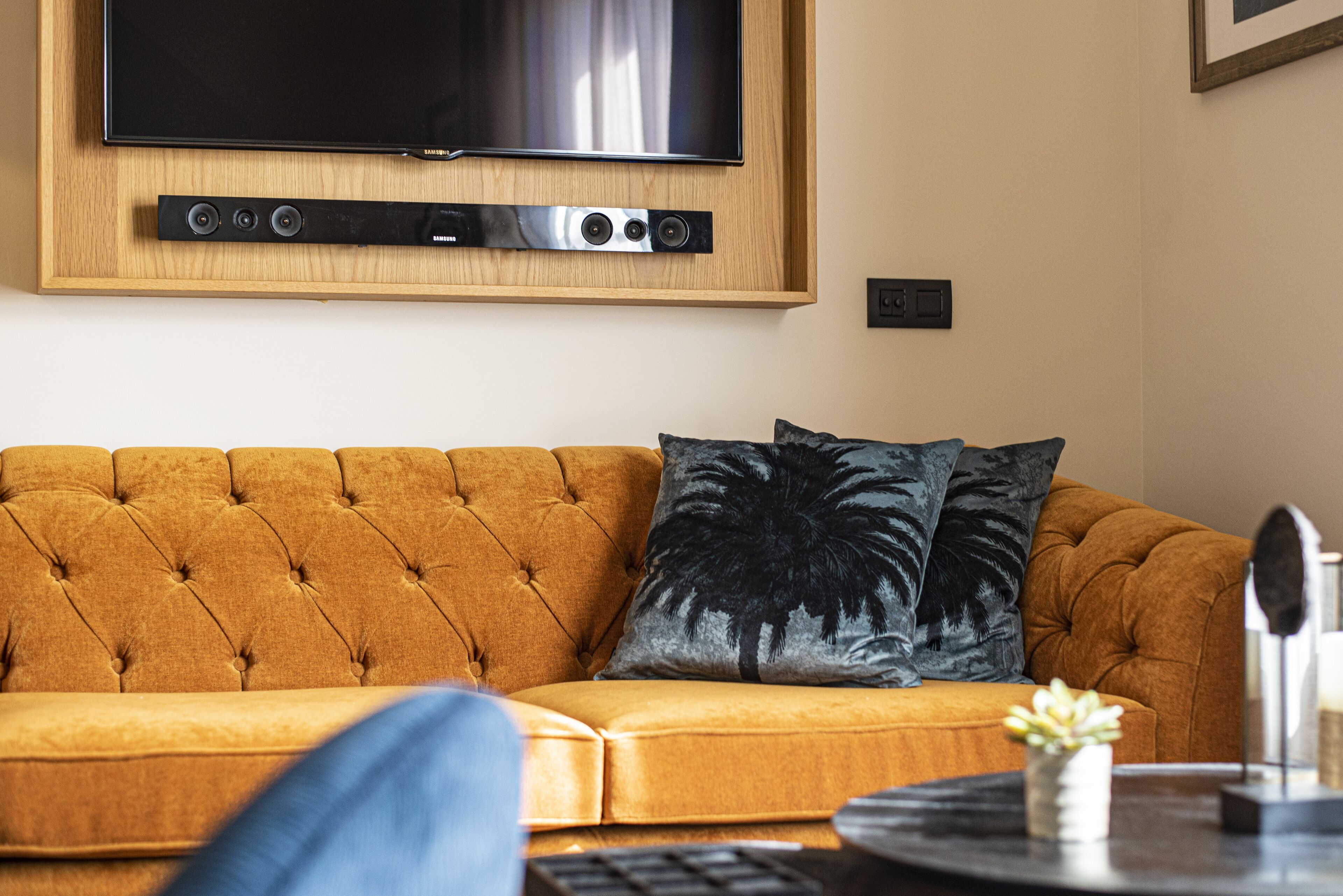 Un salon avec un canapé capitonné, un téléviseur à écran plat fixé au mur et une petite table ornée d’un objet décoratif.