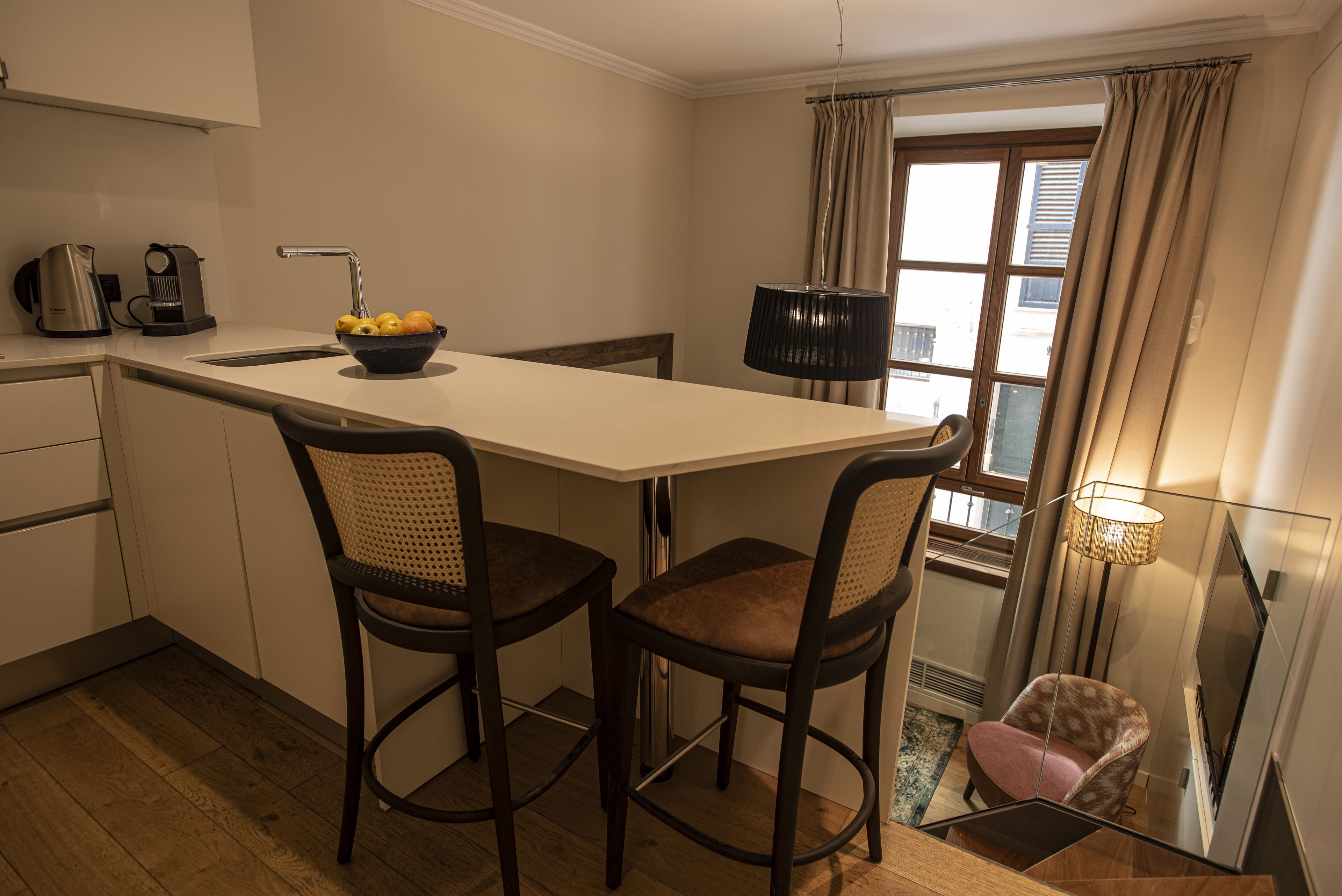 A compact kitchen with a white countertop, two bar stools, a bowl of fruit, a kettle, and a lamp.