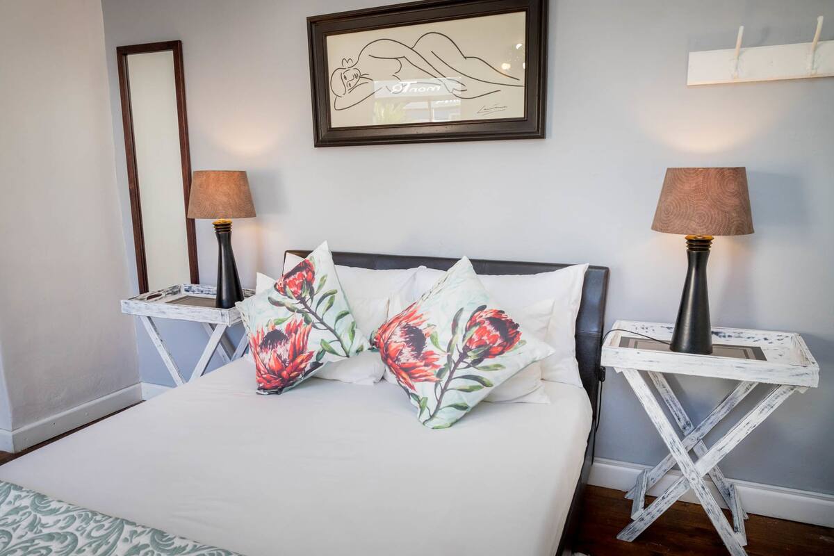 A bed with floral pillows, two bedside tables with lamps, a framed artwork on the wall, and a whiteboard on the right.