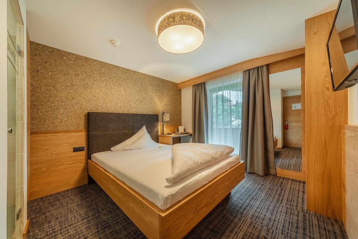 A hotel room with a large bed, wooden headboard, a bedside table with a lamp, a mirror, and a view of the outdoors through a window with curtains.