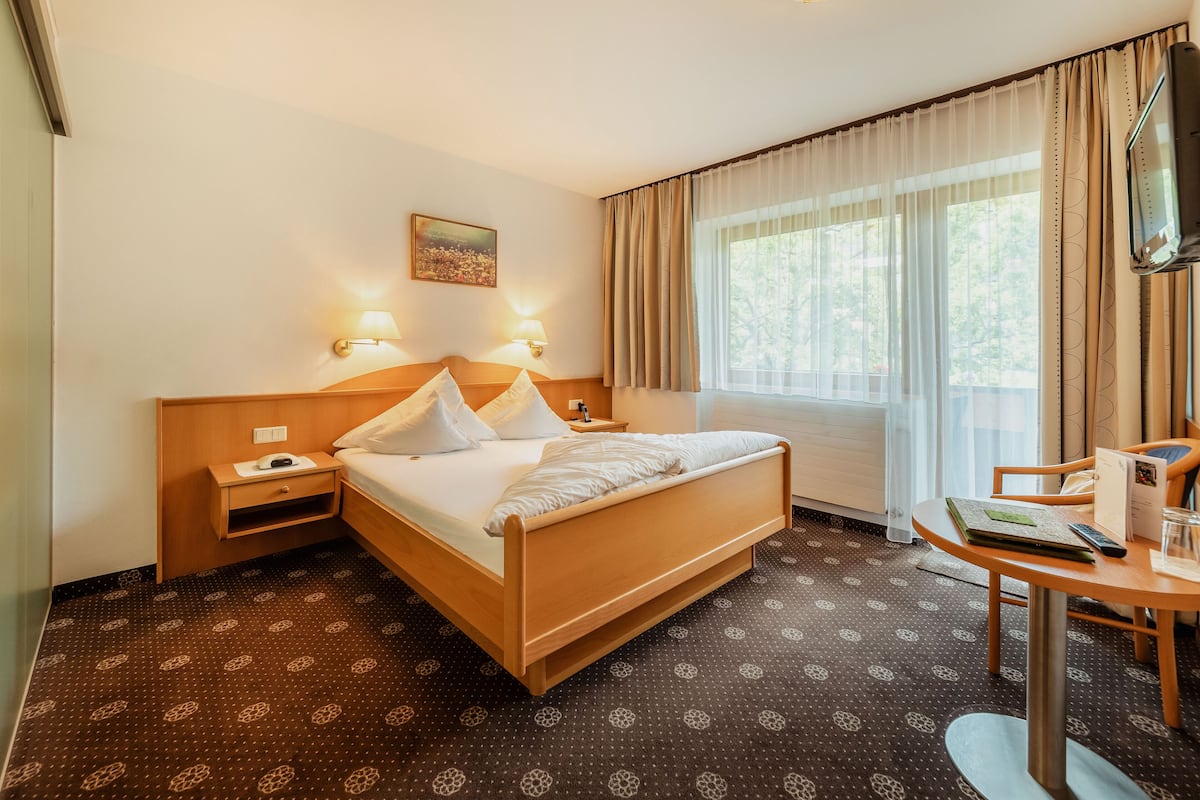 A hotel room with a wooden bed, two bedside tables with lamps, a desk with a chair, and a large window with curtains.