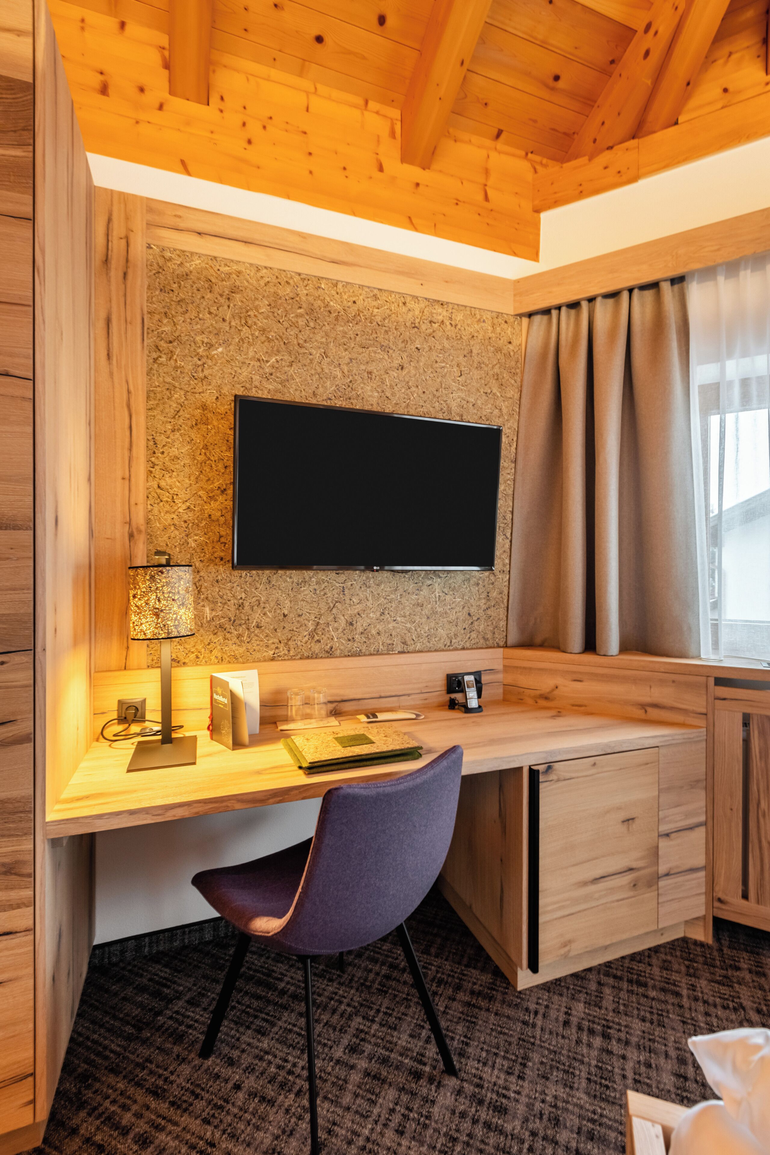 A modern room with a wooden desk, a flat-screen TV mounted on a textured wall, a lamp, and a chair.