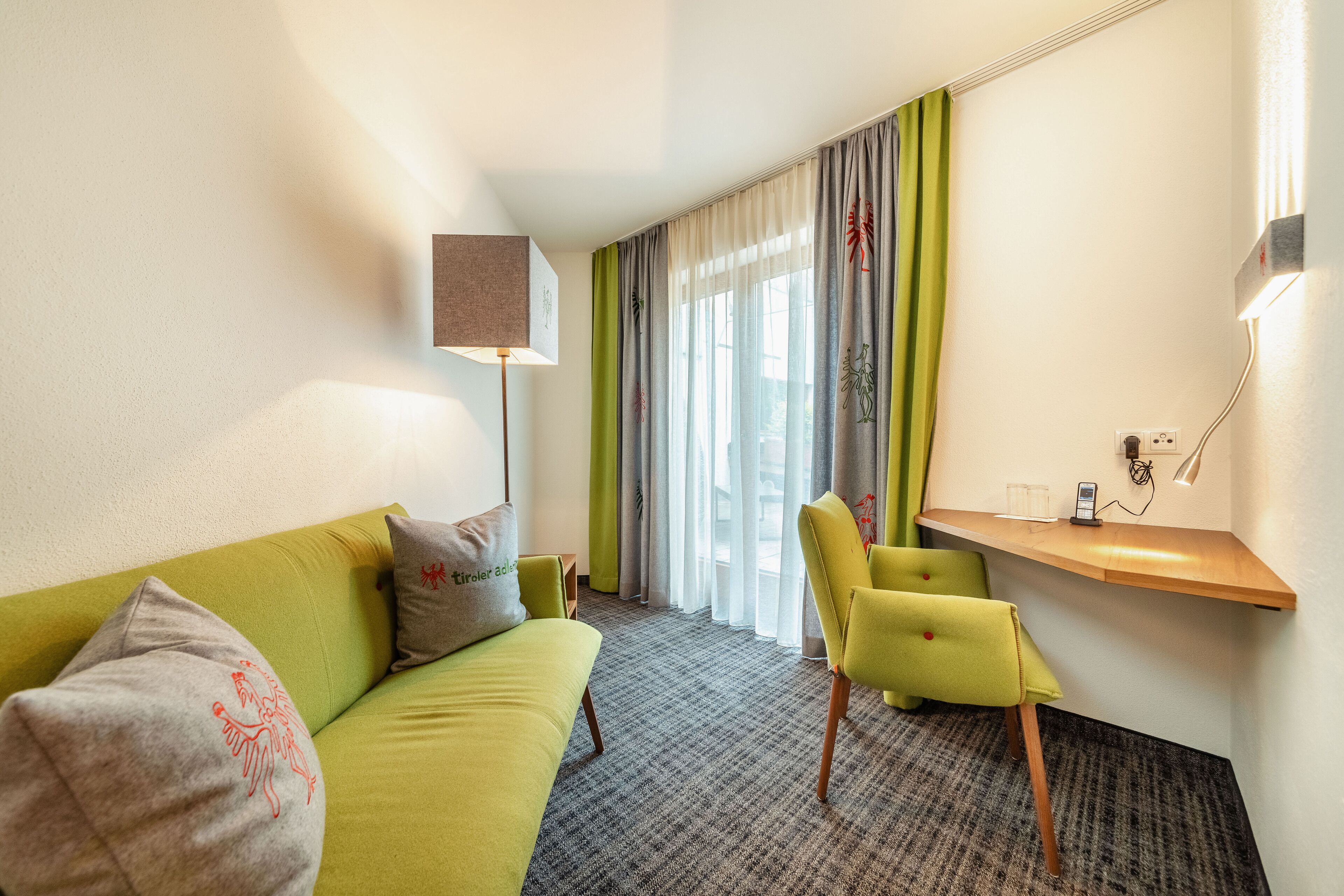 A hotel room with a green sofa, a small desk, a lamp, and a window with curtains.