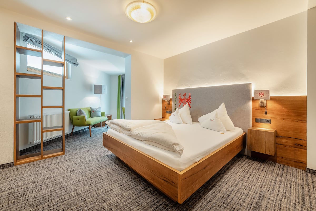 A modern hotel room with a wooden bed, a ladder, a green chair, and a view of the outdoors.