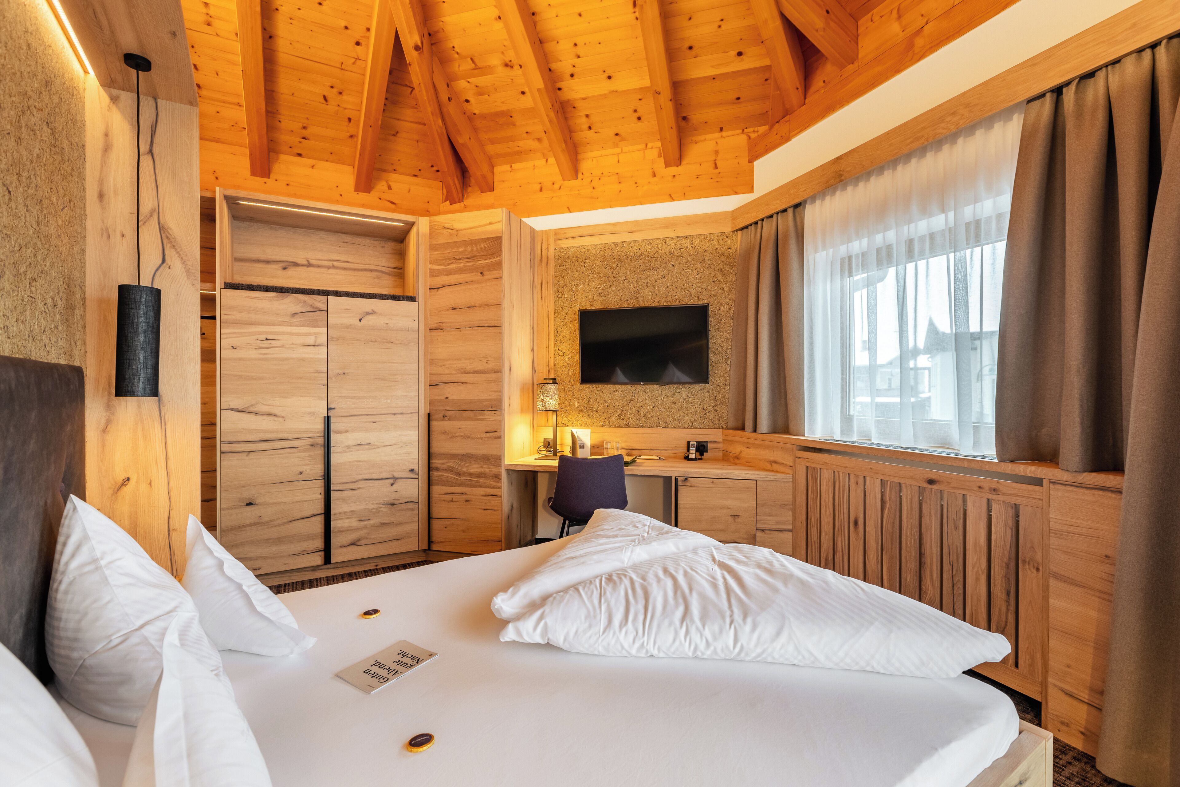 Ein Hotelzimmer mit Holzbalkendecke, einem Bett mit weißer Bettwäsche, einem Schreibtisch mit Stuhl, einem an der Wand befestigten Fernseher und einem Fenster mit Vorhängen.