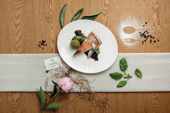 Una mesa de madera con un plato blanco que contiene una porción de tarta, una cucharada de crema y una ramita de albahaca.