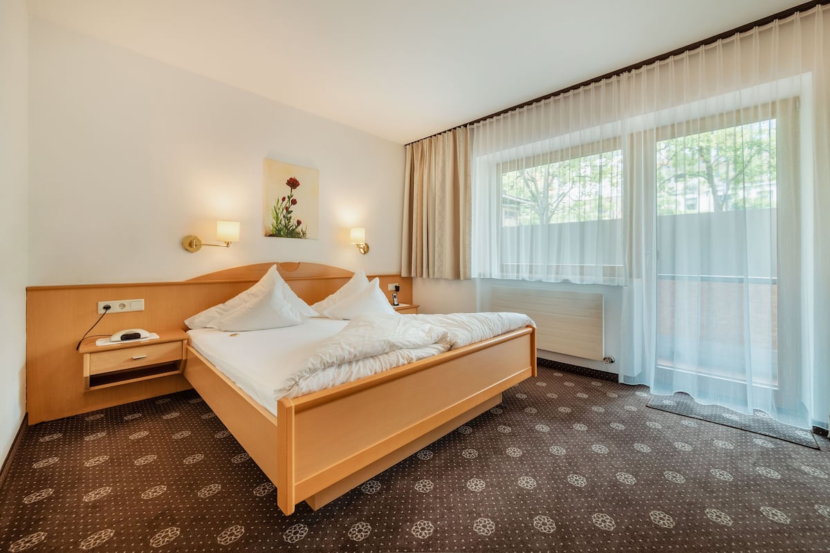 A hotel room with a wooden bed, two bedside tables, a wall-mounted lamp, and a large window with sheer curtains.