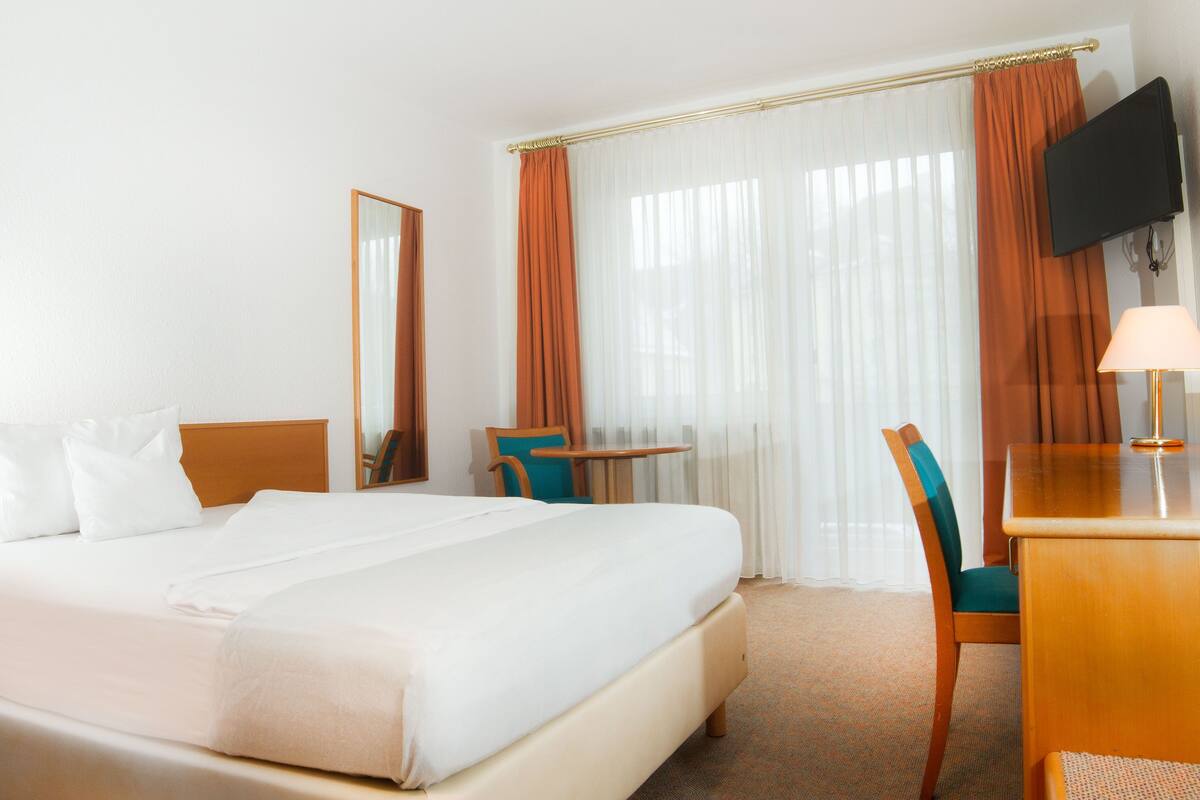 Une chambre d’hôtel avec un lit, un bureau, une chaise, une télévision et une fenêtre avec des rideaux.
