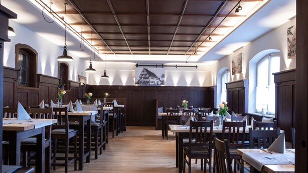 Un restaurante con mesas y sillas de madera, un techo alto con vigas a la vista y amplias ventanas que dejan entrar luz natural.