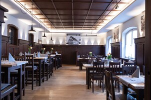 A restaurant with wooden tables and chairs, a high ceiling with exposed beams, and large windows allowing natural light.