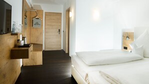 Una habitación de hotel moderna con suelo de madera, una cama con sábanas blancas, un televisor montado y un estante de madera con cafetera y vasos.
