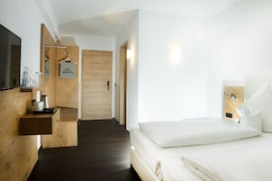 A modern hotel room with a wooden floor, a bed with white linens, a mounted TV, and a wooden shelf with a coffee maker and glasses.