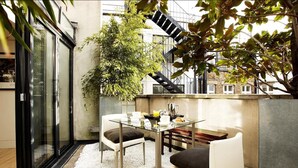 A modern outdoor dining area with a glass table and chairs, a potted plant, and a staircase in the background. - No.5 Maddox Street (London)