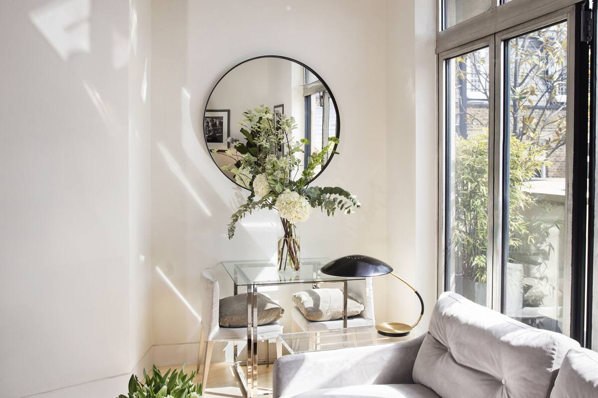 A modern living room with a sofa, a coffee table with a book, a mirror, and a window with a view of greenery.
