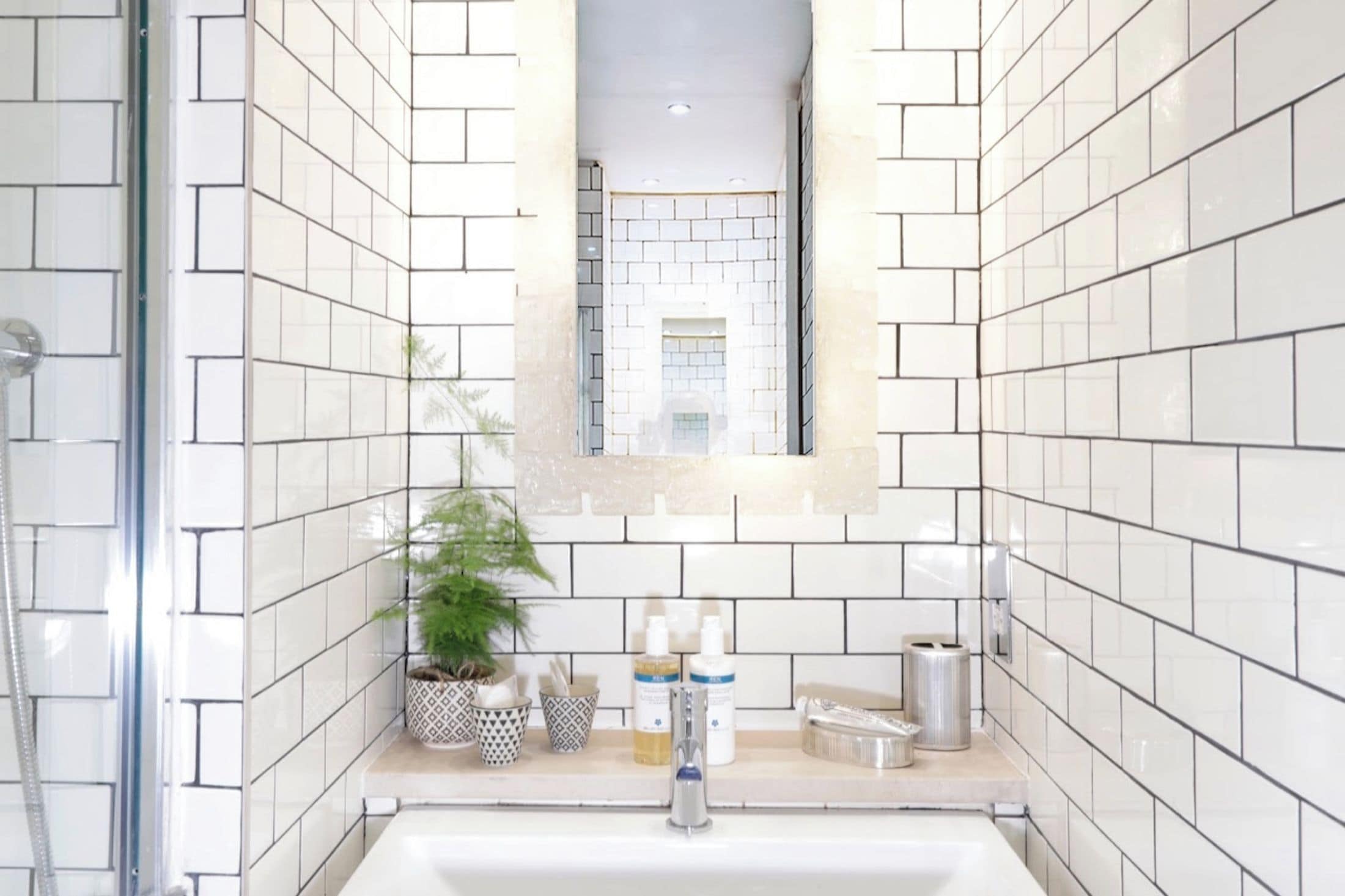A bathroom with white tiled walls, a sink, a mirror, a shower, and a potted plant.