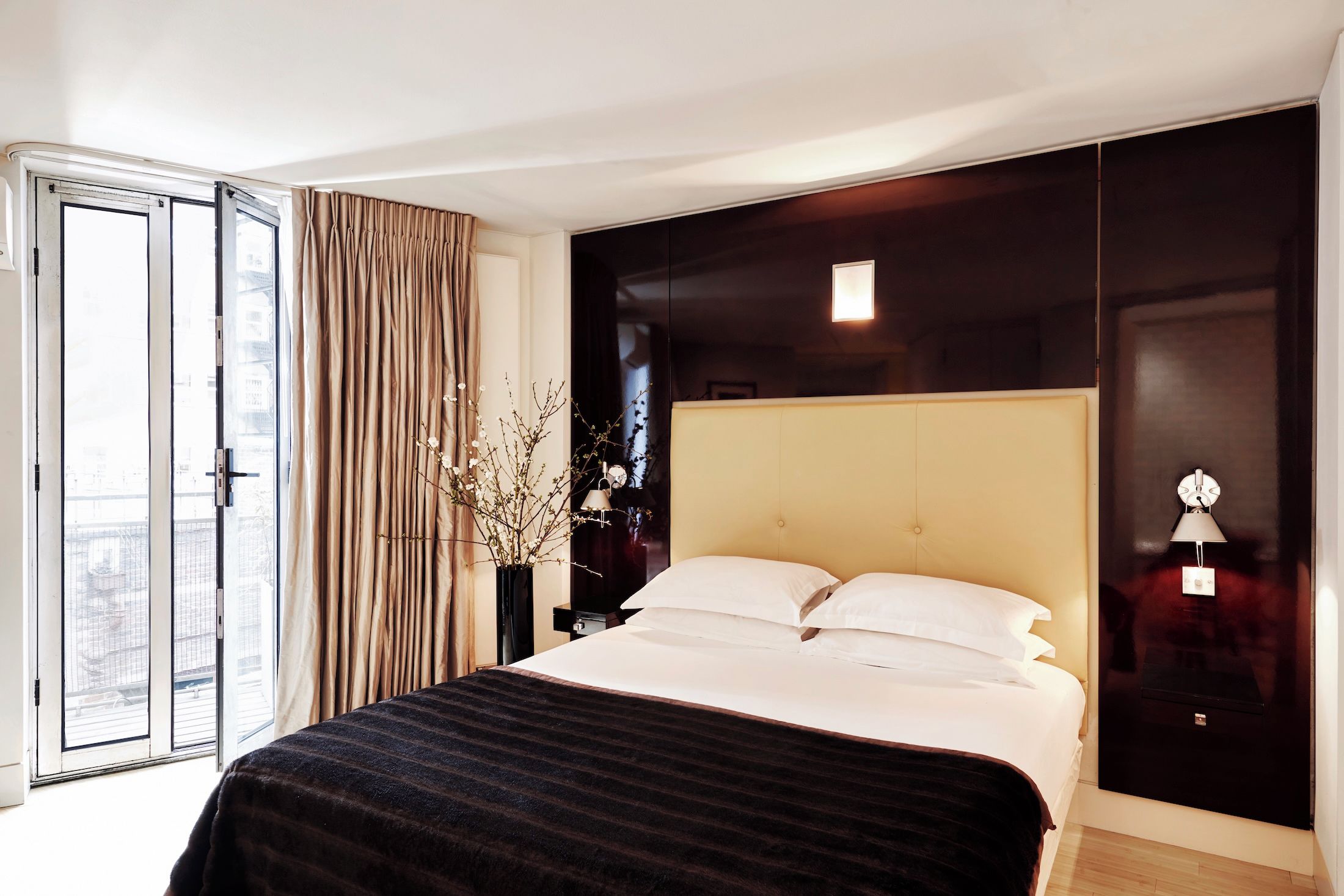 Une chambre d’hôtel avec un lit, une grande fenêtre avec des rideaux, une table de chevet avec une lampe et une armoire de couleur sombre.