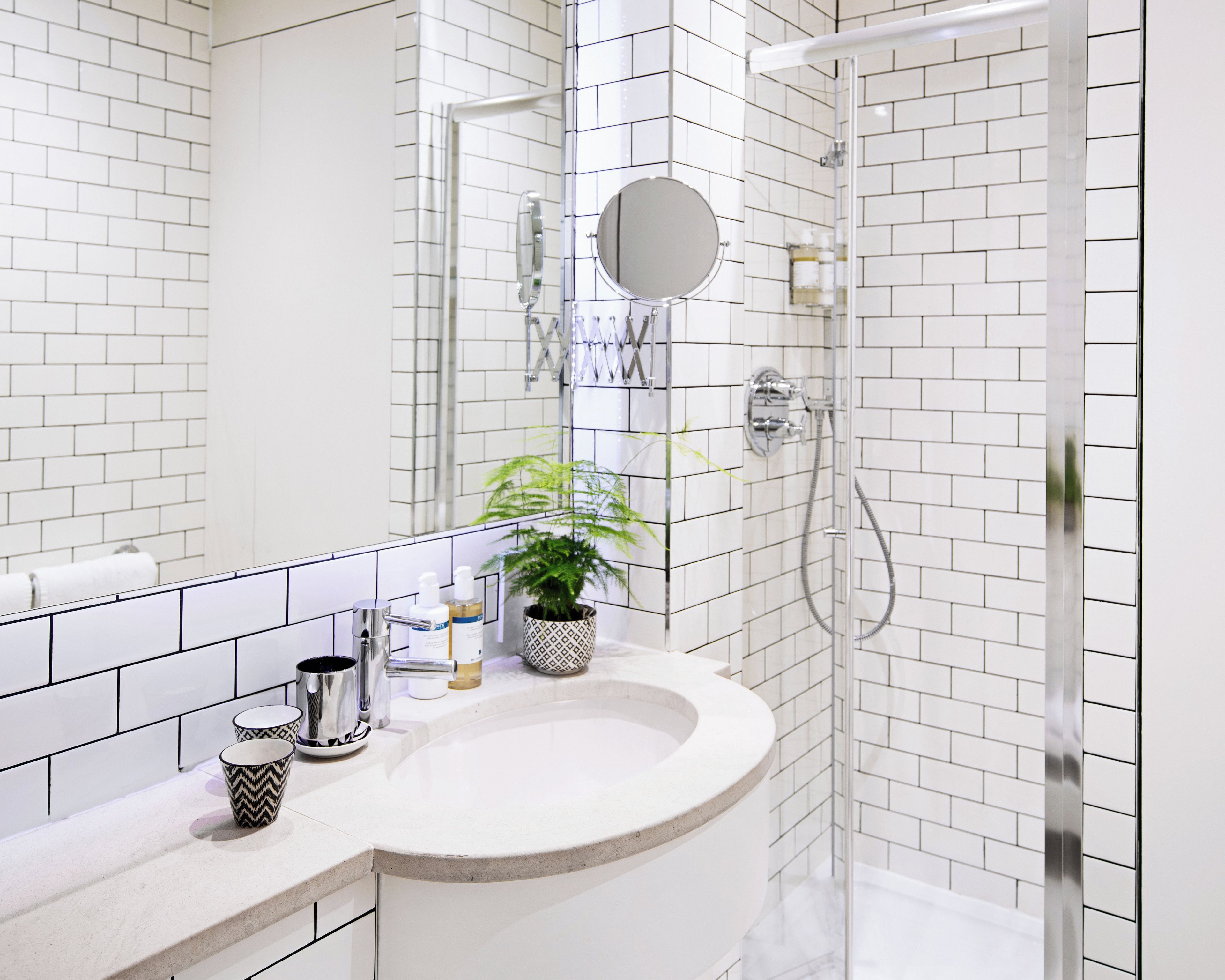 A modern bathroom with white subway tiles, a white sink, a mirror, a shower with a glass door, and a potted plant on the counter.