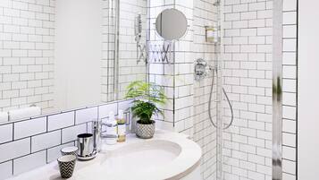 A modern bathroom with white subway tiles, a white sink, a mirror, a shower with a glass door, and a potted plant on the counter.