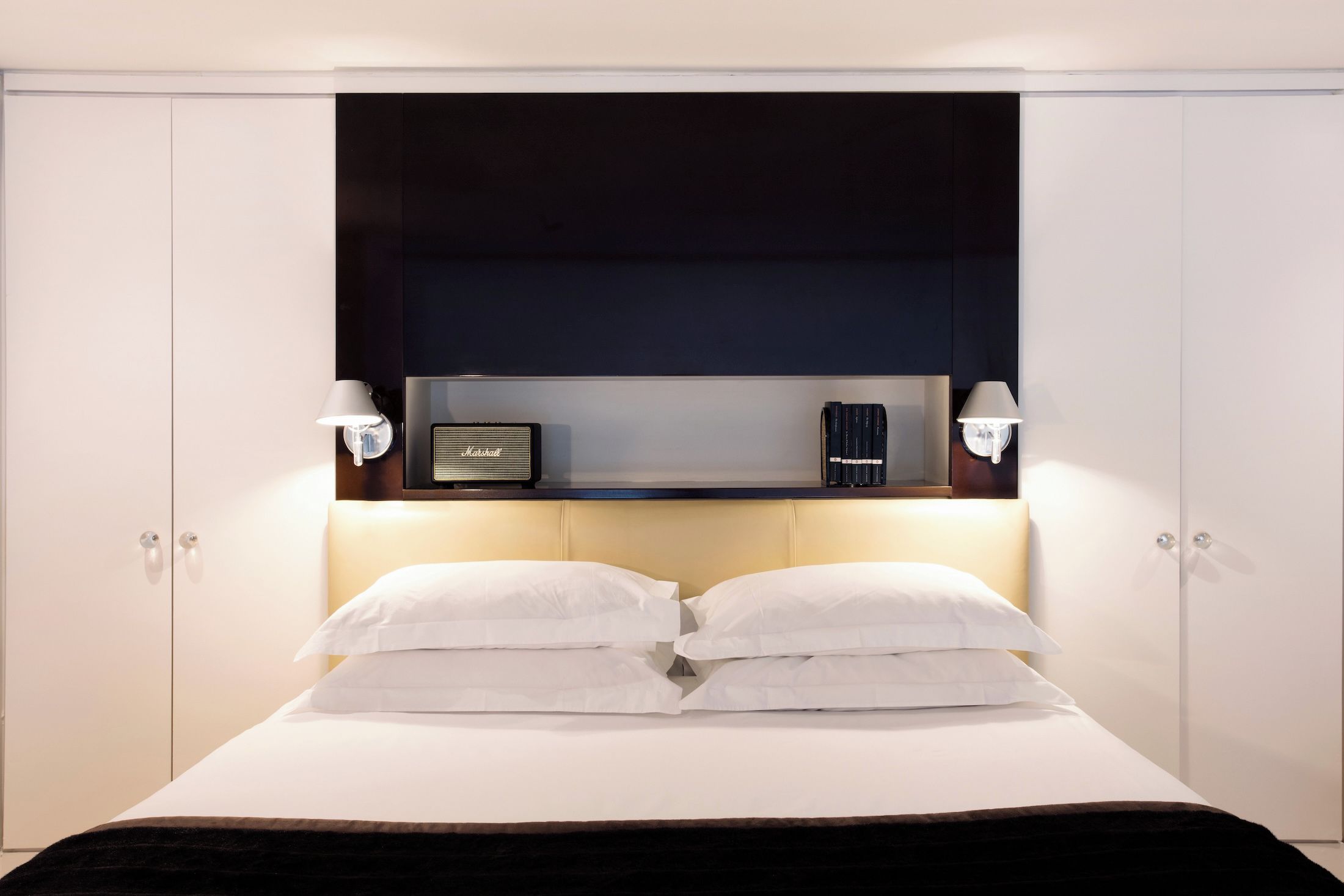A bed with white linens, a dark headboard, and two wall-mounted lamps.
