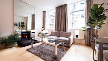 A modern living room with a sofa, a glass coffee table, a fireplace, and large windows with curtains.