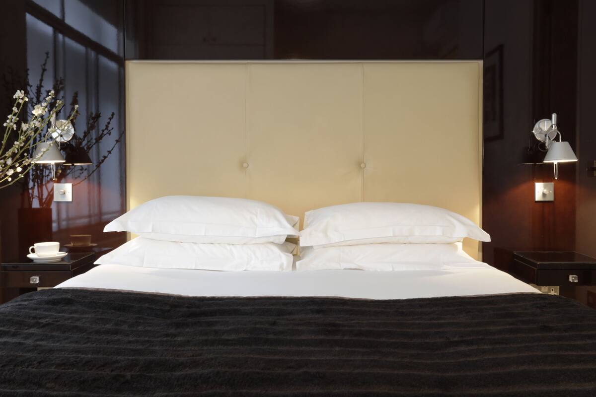A neatly made bed with white linens and a dark throw blanket, flanked by two bedside tables with lamps, a vase with flowers, and a cup on one table.