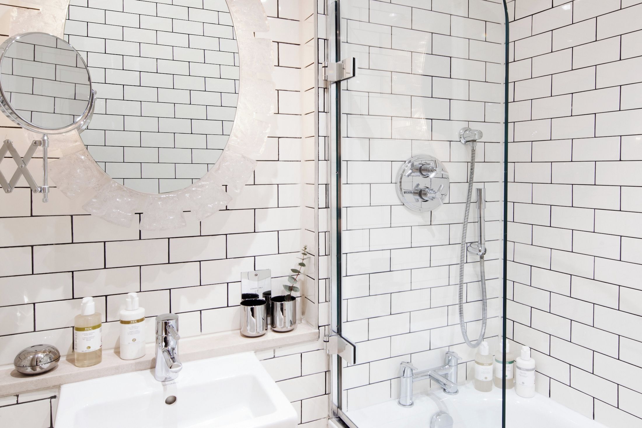 Une salle de bain avec des murs recouverts de carreaux de céramique blancs rectangulaires, une douche en verre, un miroir rond et un lavabo avec des articles de toilette.