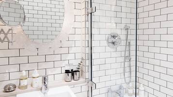 Une salle de bain avec des murs recouverts de carreaux de céramique blancs rectangulaires, une douche en verre, un miroir rond et un lavabo avec des articles de toilette.