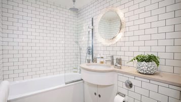 A bathroom with white subway tiles, a white bathtub, a round mirror with a decorative frame, a sink with toiletries, and a plant on the countertop.