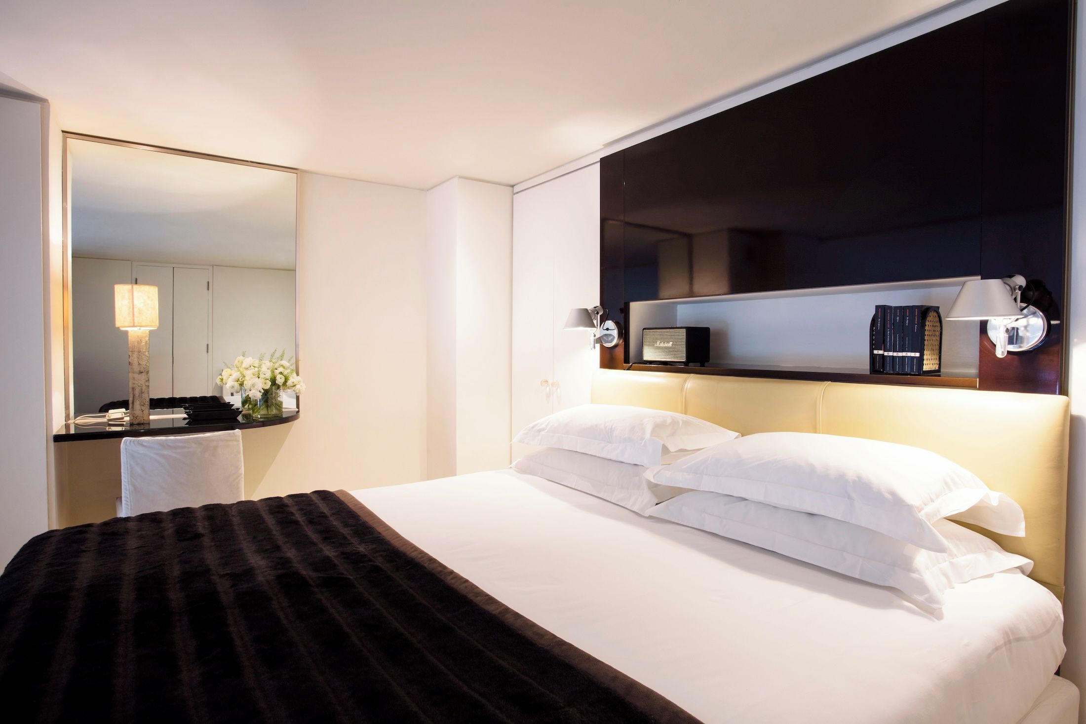 Habitación de hotel con una cama grande, una mesita de noche con lámpara, un espejo y una estantería con libros y una radio.