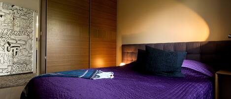 A bedroom with a purple bedspread, a wooden wardrobe, and a wall with doodle art.