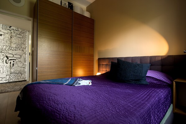 A bedroom with a purple bedspread, a wooden wardrobe, and a wall with doodle art. - King George 83 Vacation apartments (Tel Aviv)