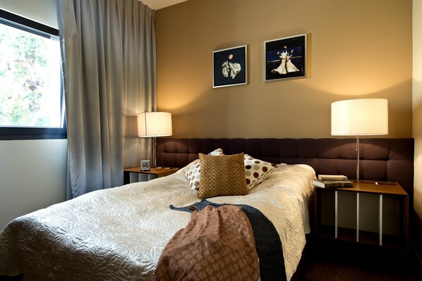 A neatly made bed with a patterned bedspread, two wall-mounted lamps, and two framed pictures on the wall.
