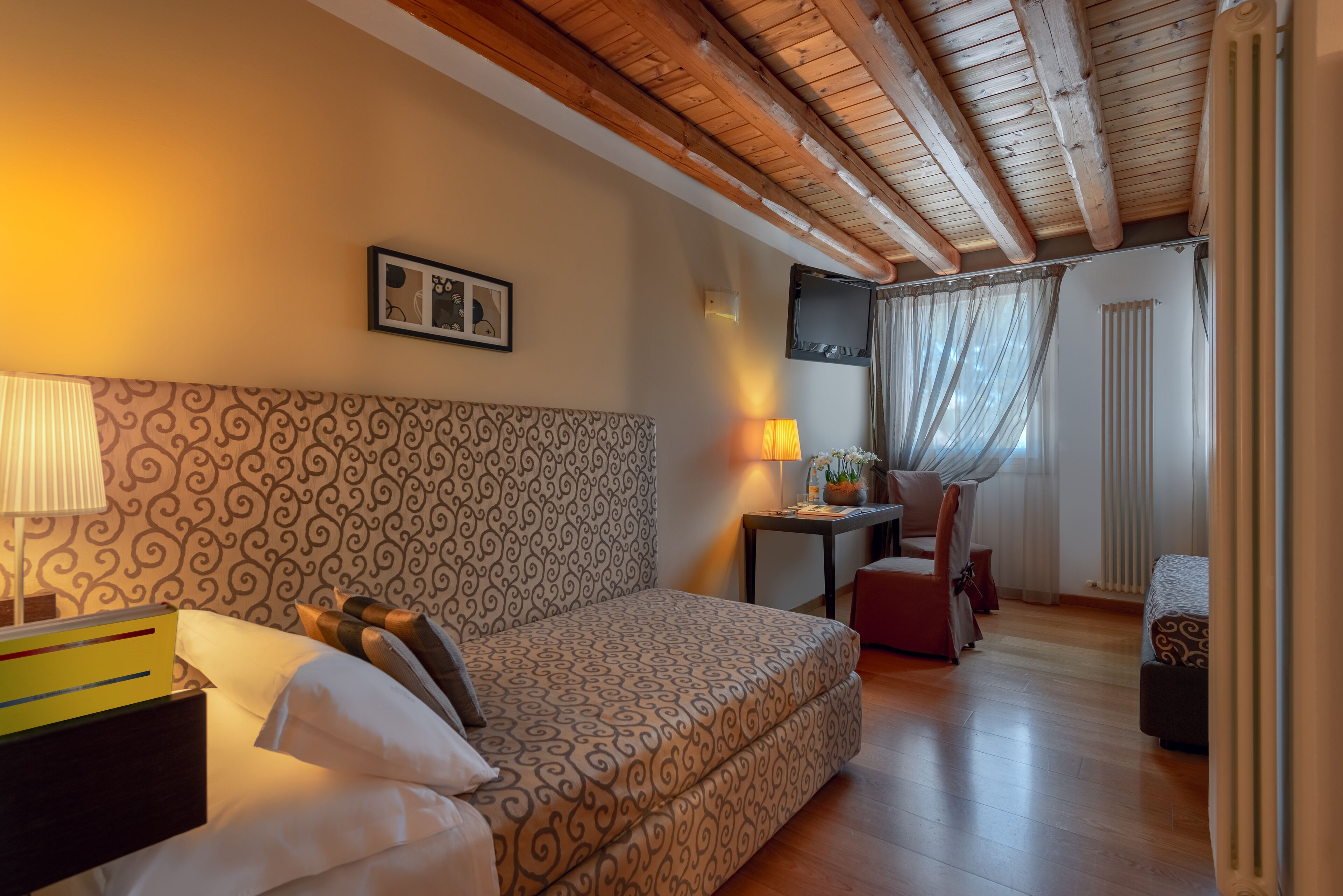 A hotel room with a patterned headboard, a bedside table with a lamp, a small desk with a chair, and a window with curtains.